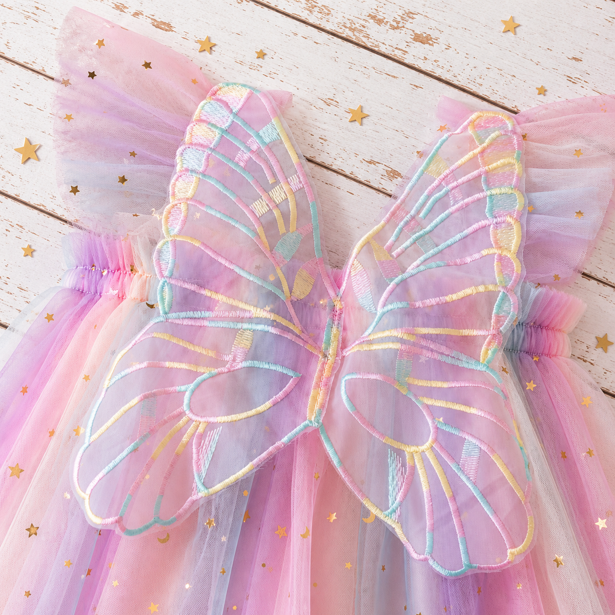 A pastel pink and lavender fairy wings costume accessory with iridescent multicolor embroidered outlines and gold star tulle, displayed on a rustic white wooden background.