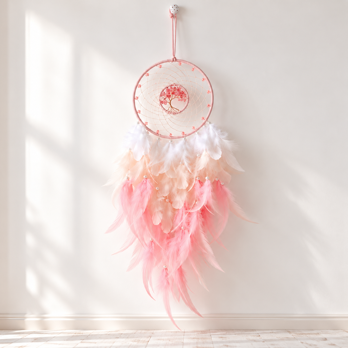 A dreamcatcher wall hanging with a pink metal hoop, white webbing, and layered feathers in white, peach, and pink, suspended against a white wall with soft window shadows.
