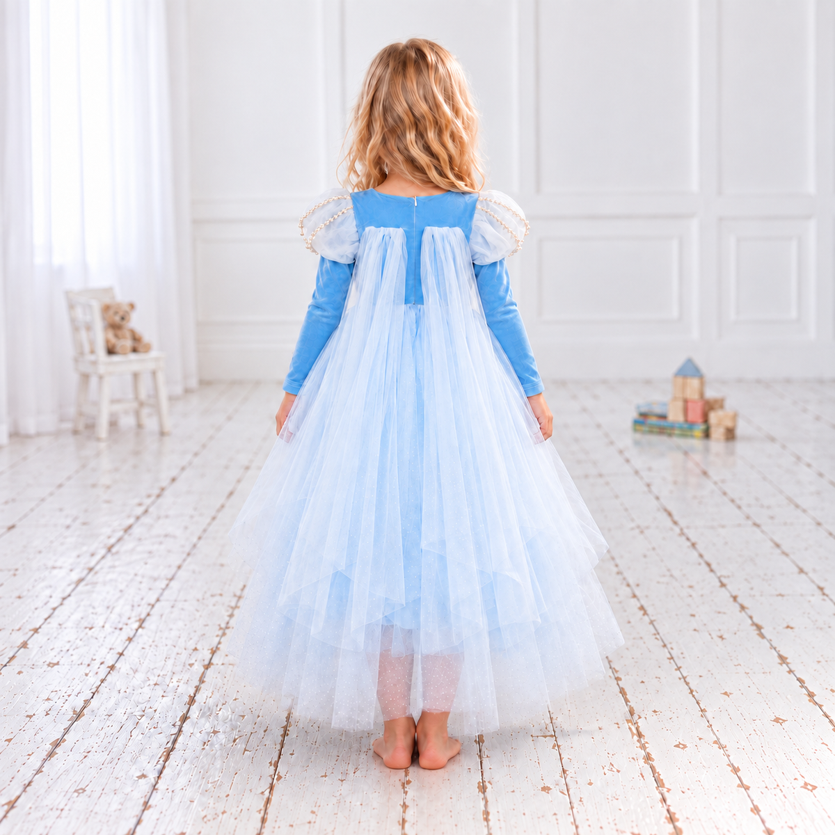  Elsa-inspired sky-blue princess dress in a white studio. The Frozen-inspired costume has a snowflake-embroidered bodice, pearl-trimmed white puff sleeves, long blue sleeves and a full skirt of fine white net over blue fabric. Her lower legs show faintly through the sheer hem.